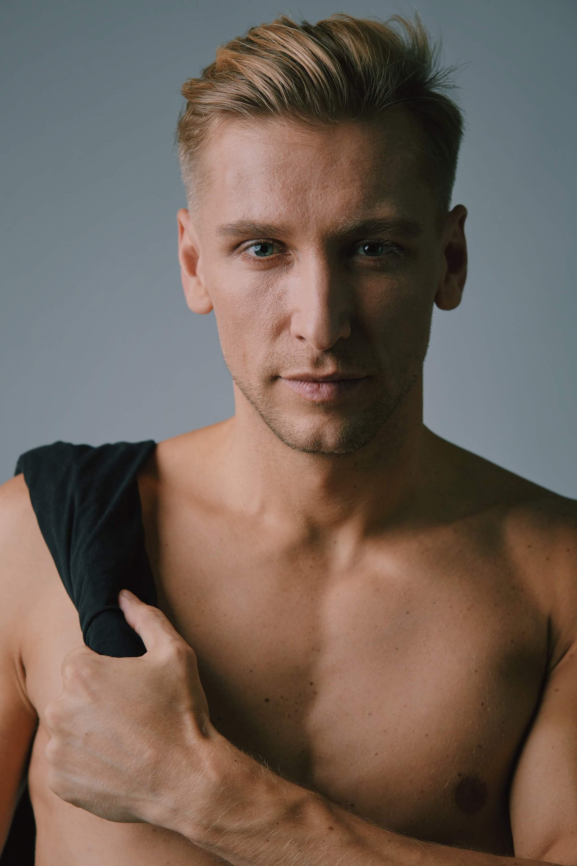 Confident shirtless man with short blond hair holding a black shirt over his shoulder, looking directly into the camera with a serious expression, in front of a neutral grey background.