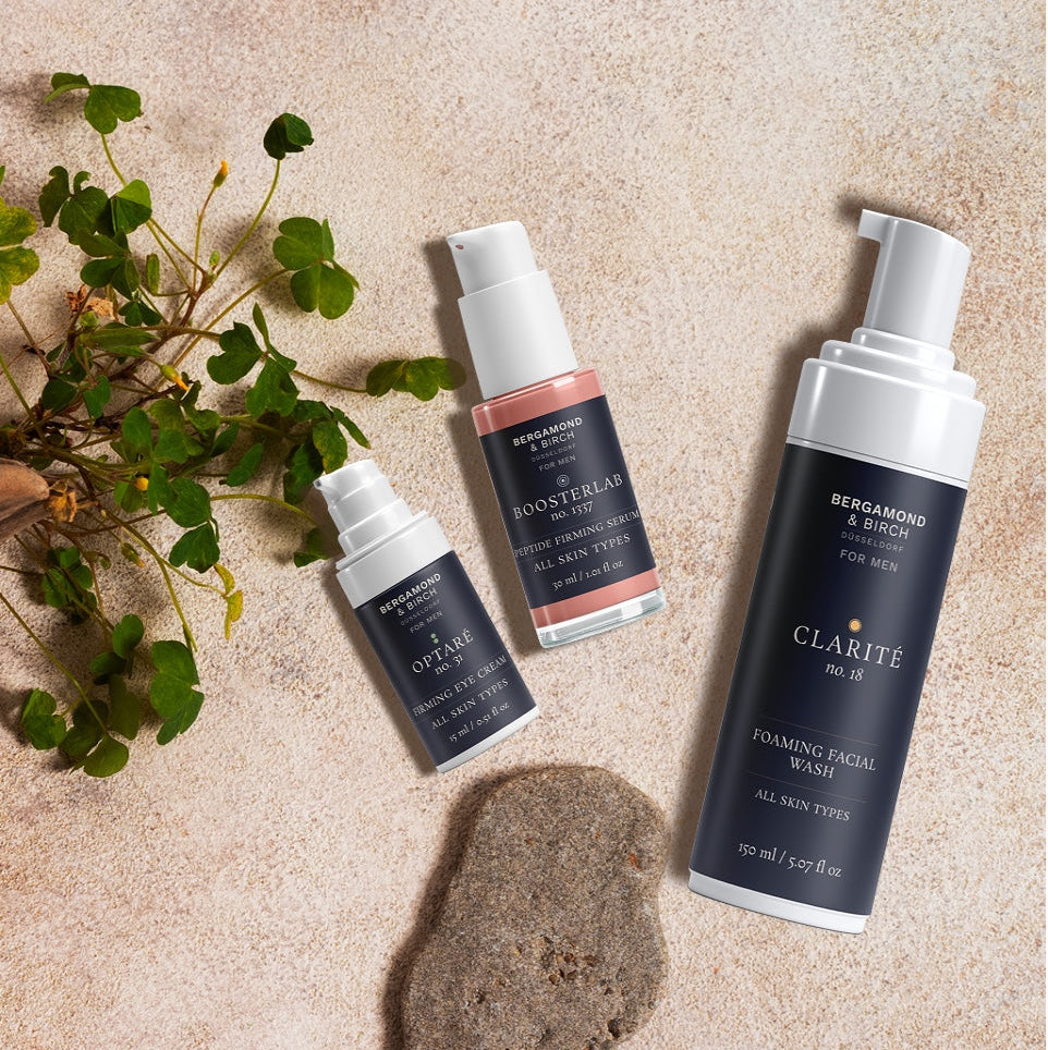 Flatlay von Bergamond & Birch Pflegeprodukten – Optaré Eye Cream, BoosterLab Peptide Firming Serum und Clarité Foaming Facial Wash – arrangiert auf einer Natursteinoberfläche mit grünen Pflanzen.