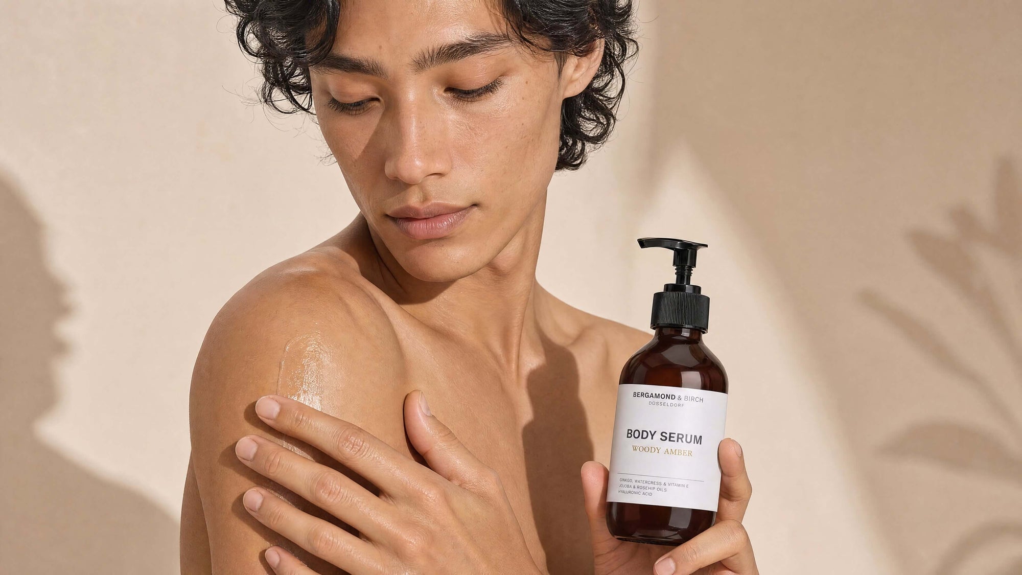 Person mit freiem Oberkörper trägt BERGAMOND & RICH Body Serum ‘Woody Amber’ auf die Schulter auf, hält die braune Pumpflasche vor beigefarbenem Hintergrund, Nahaufnahme gepflegter, glänzender Haut.
