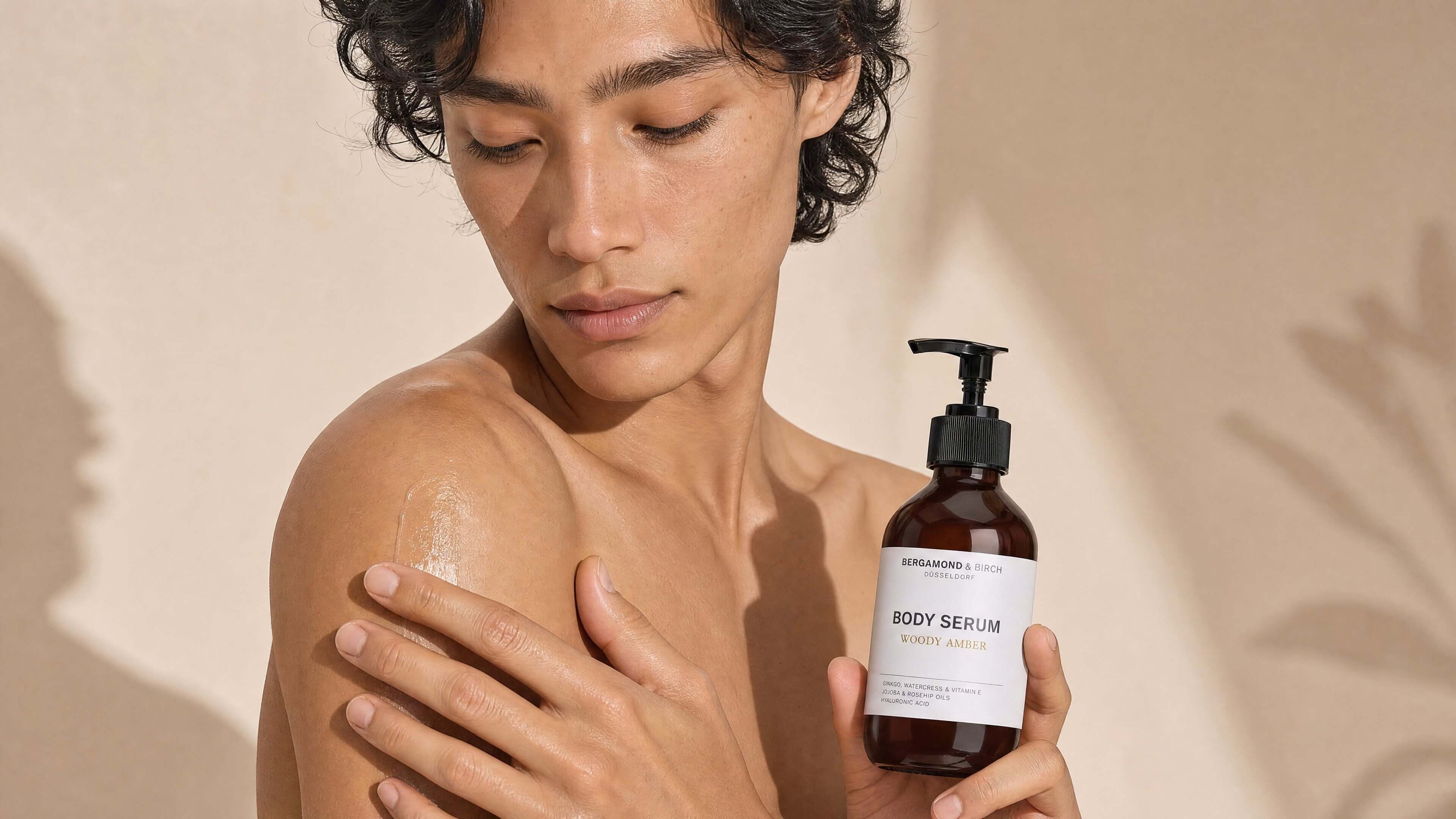 Person mit freiem Oberkörper trägt BERGAMOND & RICH Body Serum ‘Woody Amber’ auf die Schulter auf, hält die braune Pumpflasche vor beigefarbenem Hintergrund, Nahaufnahme gepflegter, glänzender Haut.