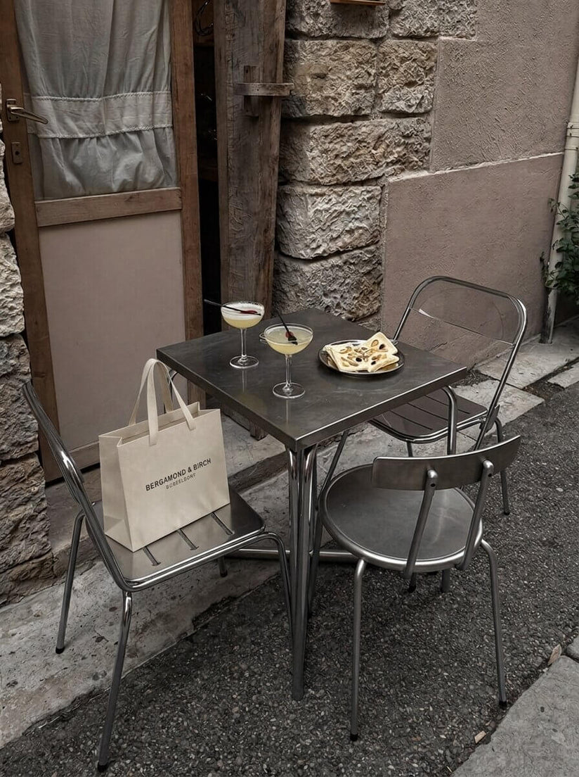 Kleiner Metalltisch vor einer rustikalen Hausfassade mit zwei Cocktailgläsern, Gebäck und BERGAMOND & RICH Shopping-Bag auf dem Stuhl, stimmungsvolle Szene eines Café-Besuchs mit hochwertiger Hautpflege-Einkaufstasche.