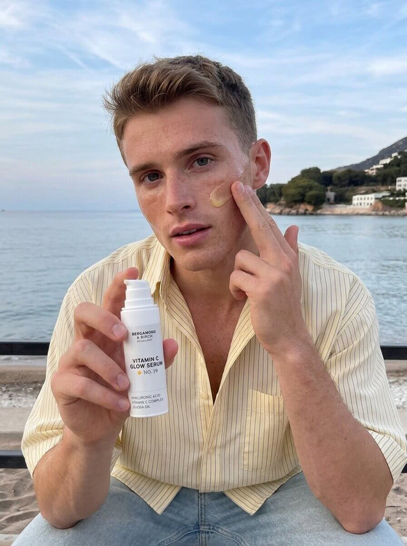 Mann sitzt am Strand und trägt BERGAMOND & RICH Vitamin C Glow Serum No. 19 auf seine Wange auf, hält die weiße Serumflasche vor Meer und Küstenlandschaft – zeigt tägliche Gesichtspflege mit Glow-Effekt im Urlaub.