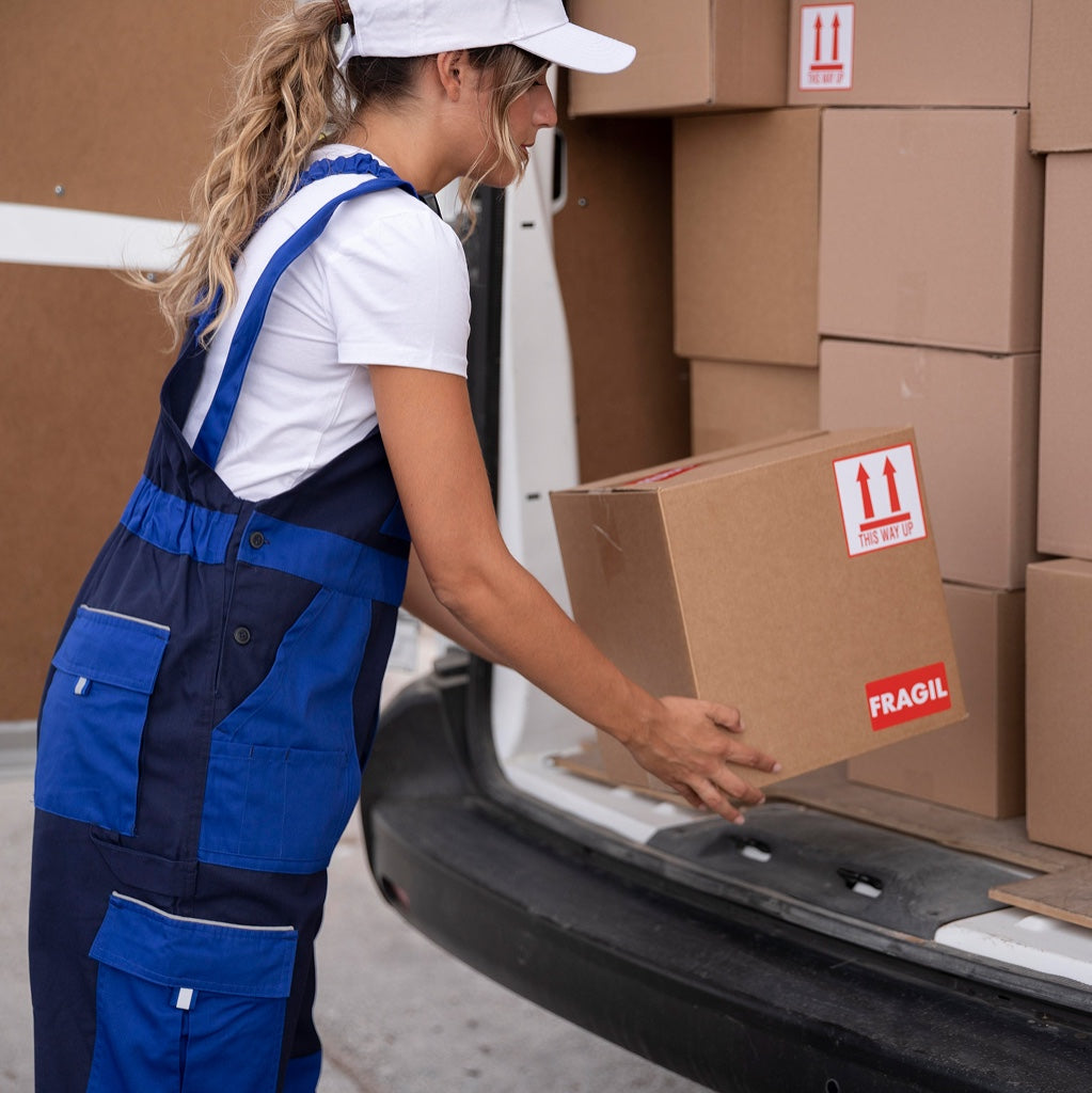 Person in Arbeitskleidung hebt einen Karton mit Fragile-Aufkleber in einen Lieferwagen voller Paketboxen.