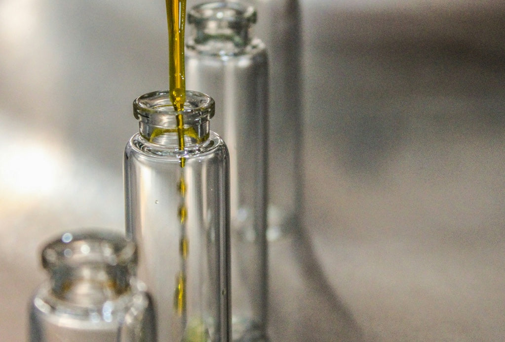 Nahaufnahme eines Abfüllprozesses, bei dem eine Pipette goldenes Öl in eine Reihe transparenter Glasfläschchen füllt.