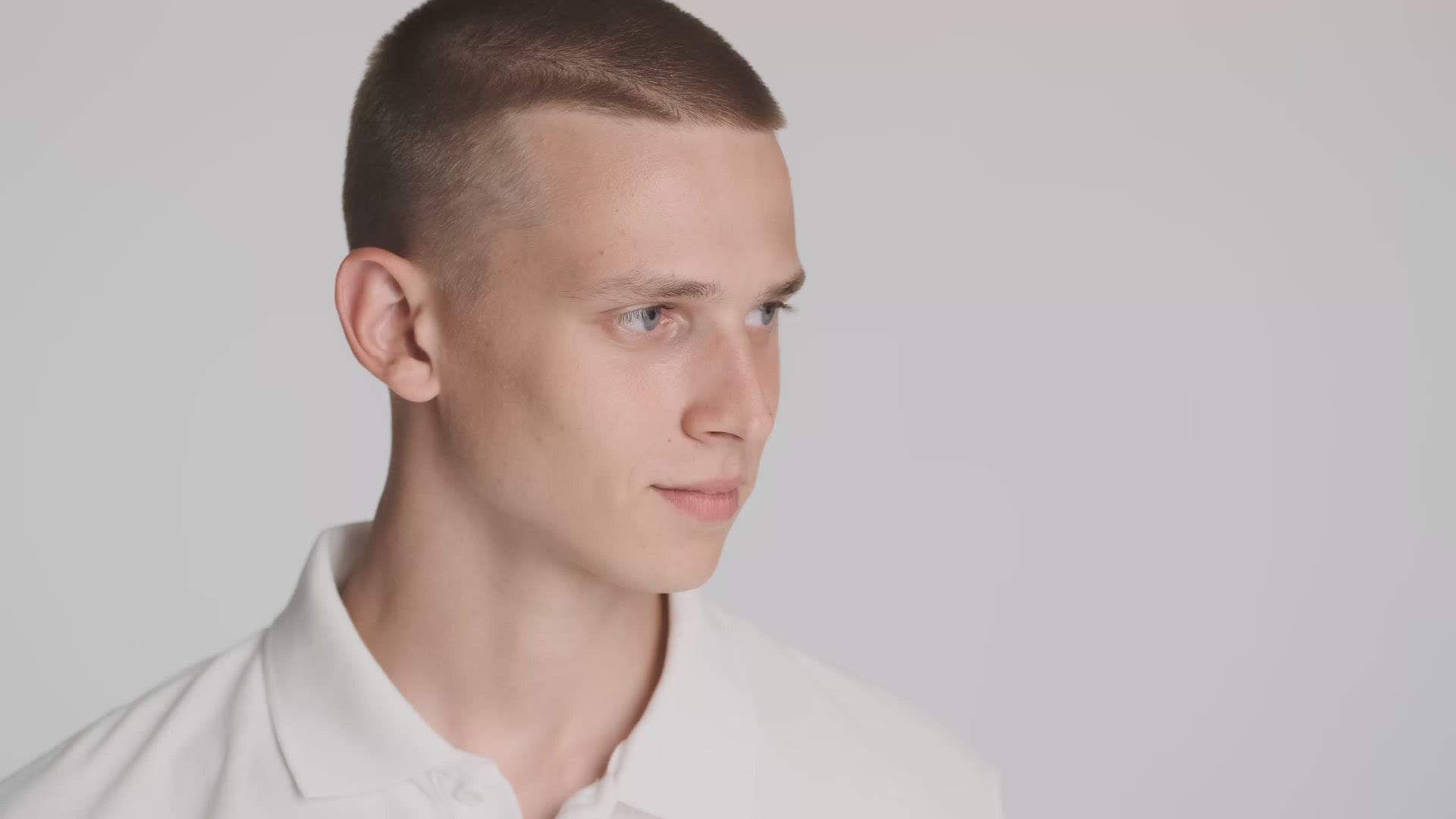 A young man with short hair and clear skin smiling confidently in front of a light grey background. He wears a white polo shirt, evoking a clean, minimal, and fresh aesthetic – ideal for skincare or wellness branding.