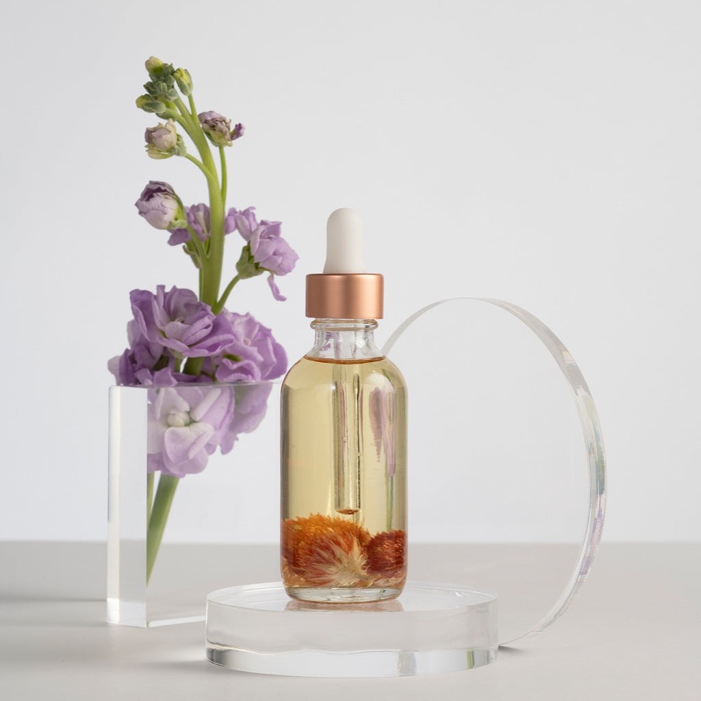 Glasflasche mit goldenem Serum und eingelegten Blüten, arrangiert vor einer lila Blume auf hellem Hintergrund